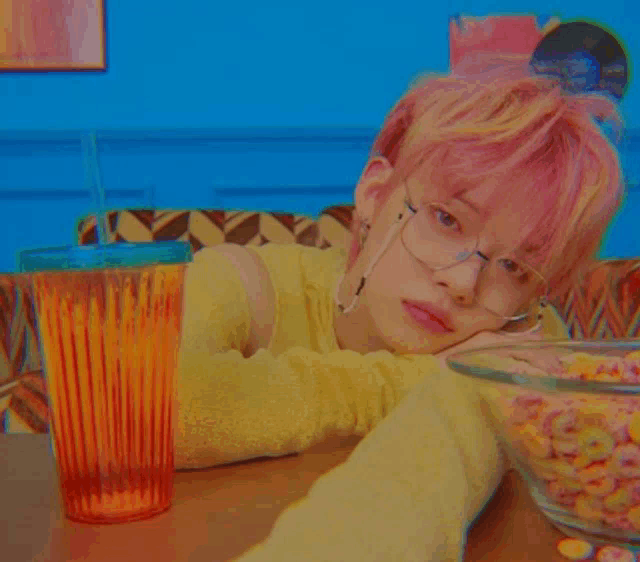 a girl with pink hair and glasses is sitting at a table with a bowl of cereal and a glass of orange juice .