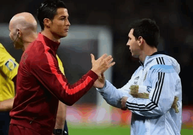 two soccer players , cristiano ronaldo and messi , shake hands on a soccer field .