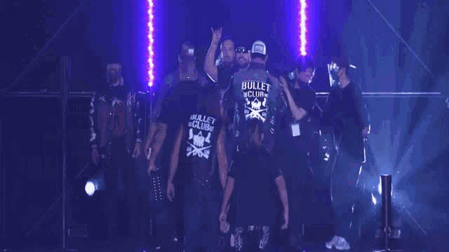 a group of men are standing in a dark room and one of them is wearing a shirt that says bullet club