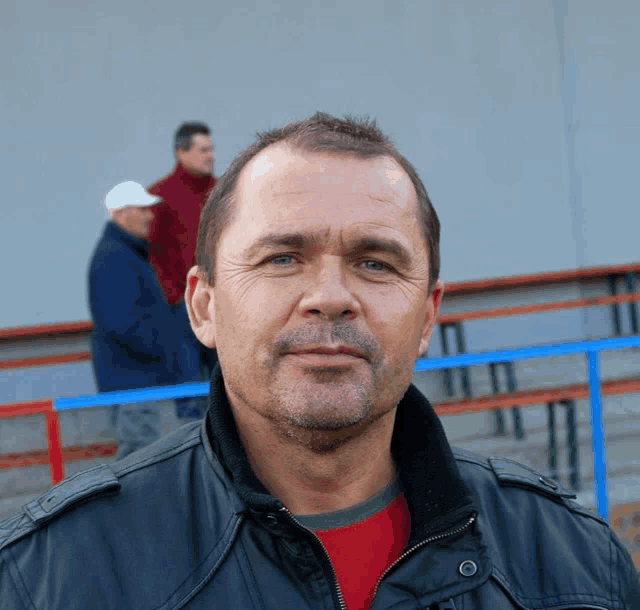 a man in a black jacket and red shirt stands in front of a blue railing