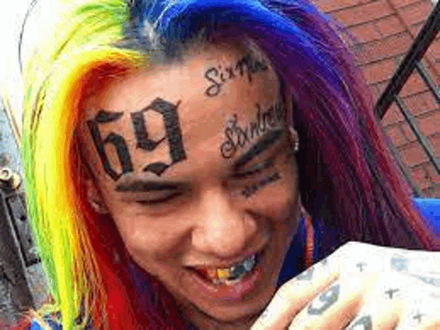 a man with rainbow hair and a tattoo on his face is holding a cell phone .