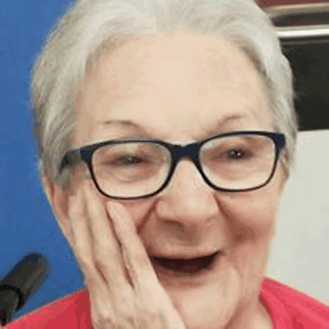 an elderly woman wearing glasses and a red shirt is smiling and touching her face .