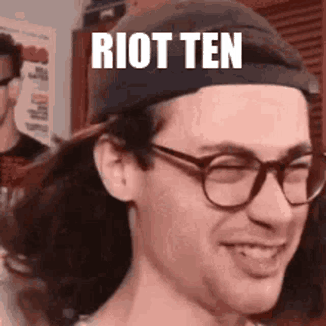 a man wearing glasses and a hat is smiling and says riot ten .