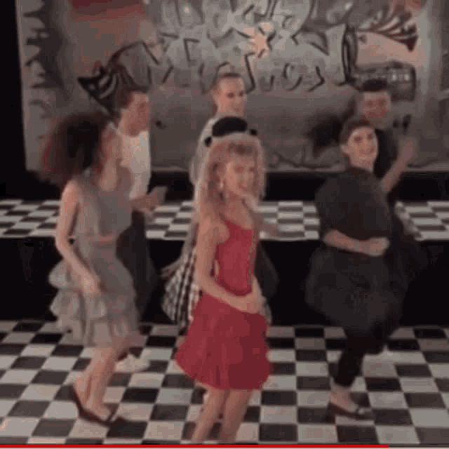 a group of people are dancing on a checkered floor and one of them is wearing a red dress