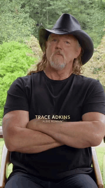 a man wearing a black shirt that says trace adkins is sitting in a chair with his arms crossed