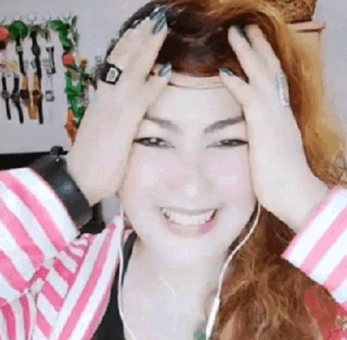 a woman in a pink and white striped shirt is smiling and holding her head with her hands .