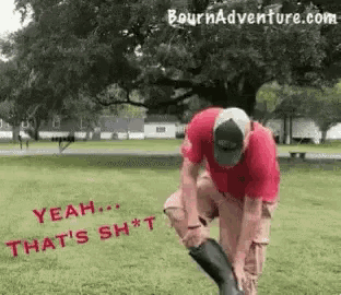 a man wearing a red shirt and a hat is putting on a pair of rubber boots in a park .