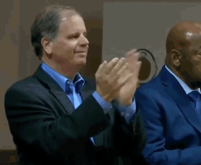 a man in a suit applauds while another man in a suit looks on