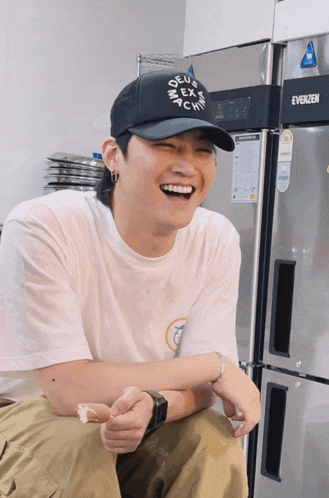 a man wearing a white shirt and a black hat is sitting in front of a evergreen refrigerator