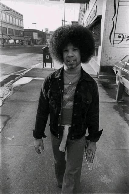 a black and white photo of a man walking down a sidewalk in front of a mailbox with the letter d on it