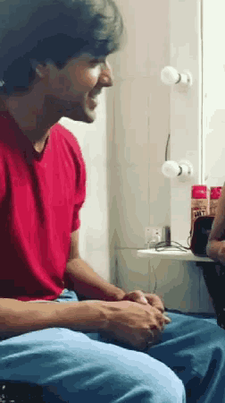 a man in a red shirt sits in front of a mirror with a bottle of salt on the counter