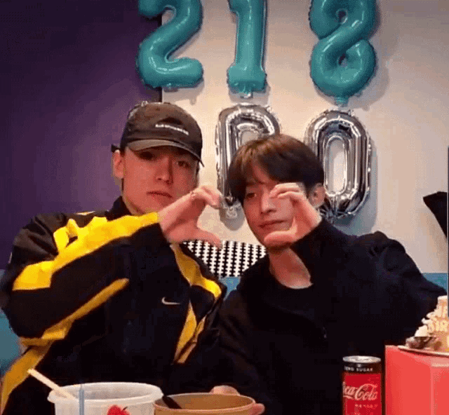 two young men are making a heart shape with their hands while sitting at a table with a can of coca cola .