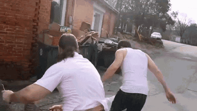 two men are running down a street in front of a brick building . one of the men is wearing a white tank top .