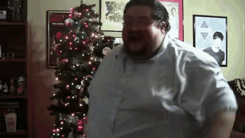 a very fat man is standing in front of a christmas tree .