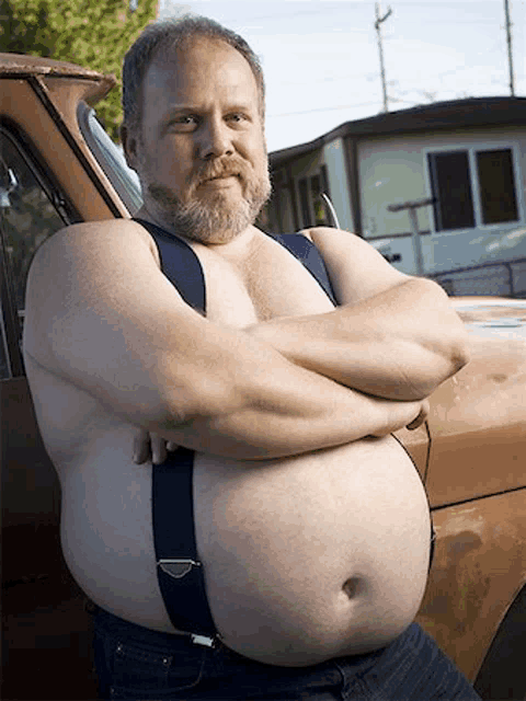 a shirtless man with a beard and suspenders is standing next to a truck .