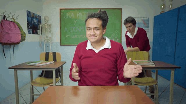 a boy in a red sweater sits at a desk in front of a green board that says una paja y a dorm