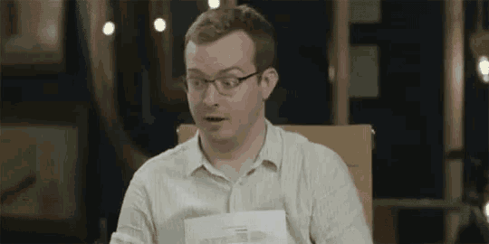 a man wearing glasses is sitting at a table holding a piece of paper and making a funny face .