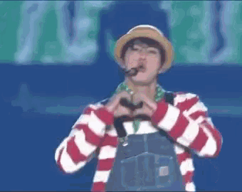 a man wearing a hat and overalls is making a heart shape with his hands while singing into a microphone .