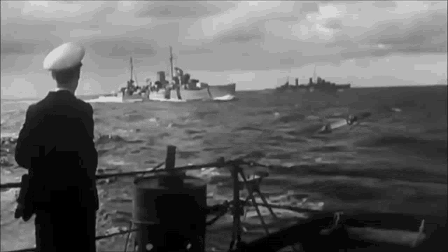 a black and white photo of a man standing on a boat looking at a large ship in the ocean