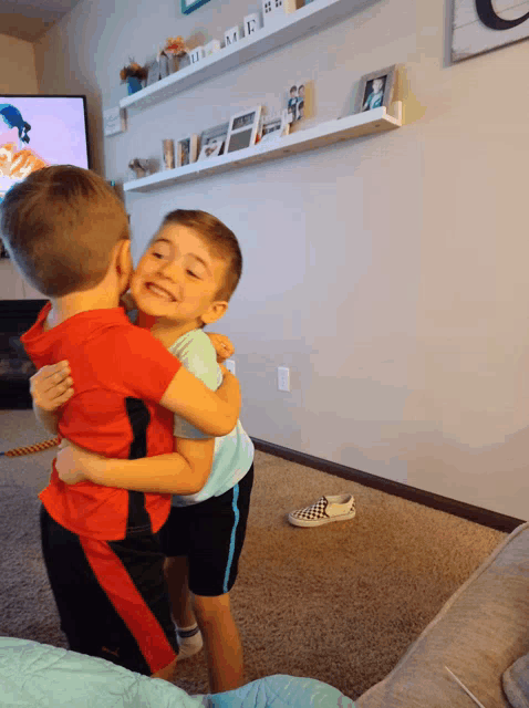 a boy in a red shirt is hugging another boy in a blue shirt in a living room