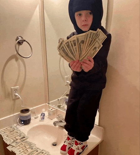 a young boy holds a fan of 20 dollar bills in front of a bathroom sink