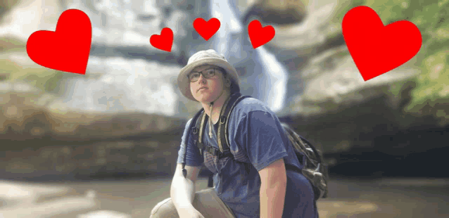 a man wearing a hat and glasses stands in front of a waterfall with red hearts above him