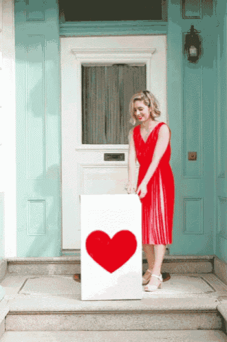 a woman in a red dress holds a white box with a red heart on it in front of a blue door