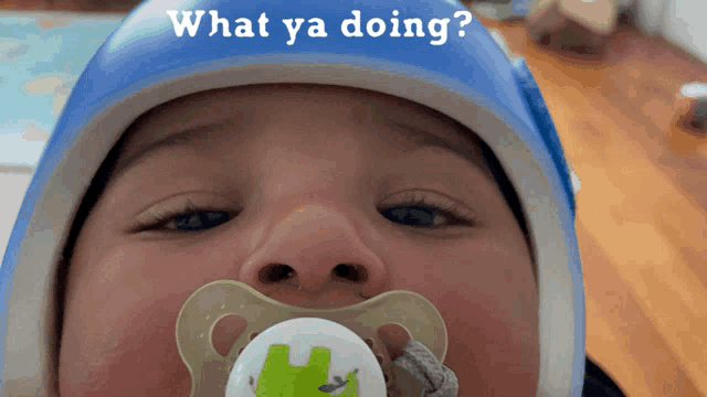 a baby with a pacifier in his mouth is wearing a blue helmet with the words what ya doing on it