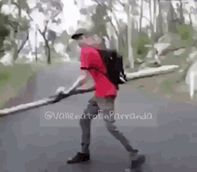 a man in a red shirt is walking down a road with a backpack and holding a large stick .