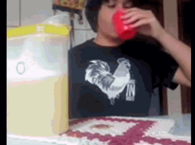 a man wearing a black shirt with a rooster on it drinking from a red cup .