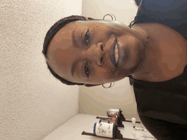 a woman with purple lipstick and hoop earrings smiles in front of a wall with a shelf of pills on it