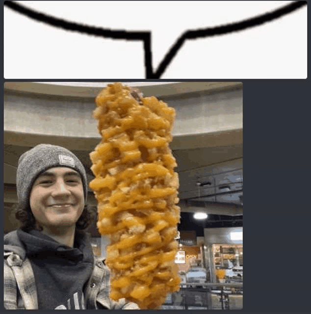 a man in a beanie is holding a very long corn dog with cheese .