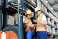 a man is driving a forklift in a warehouse while a man looks at a tablet .
