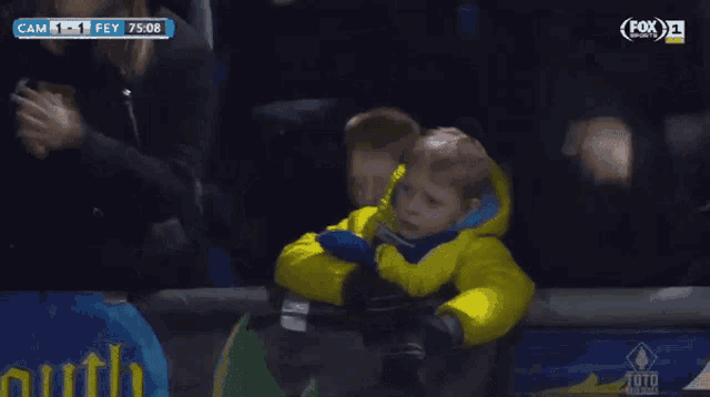 a little boy in a yellow jacket sits in a stadium watching a soccer game with fox on the screen