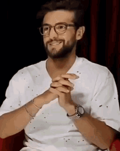 a man with a beard wearing glasses and a watch is sitting in a chair with his hands folded .