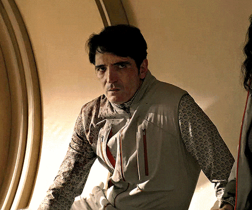 a man wearing a grey vest and a patterned shirt looks at the camera