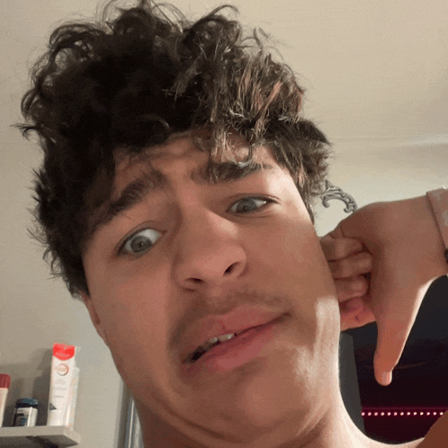 a young man with curly hair is making a funny face with his hand in his ear