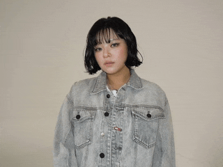 a woman wearing a grey denim jacket with black buttons looks at the camera