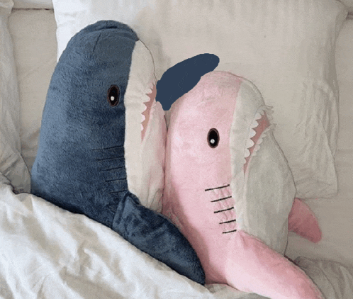 a blue and a pink stuffed shark are laying on a bed