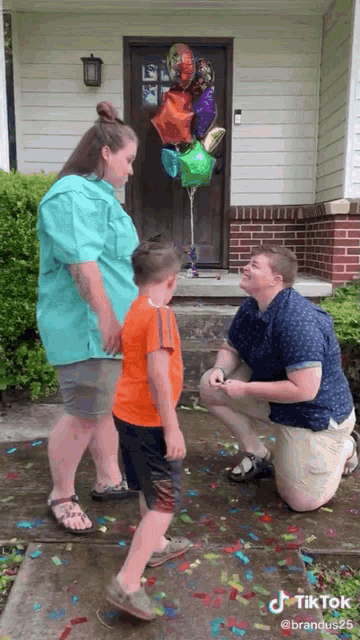 a man is kneeling down to propose to a woman and a boy in front of a house with balloons and confetti .