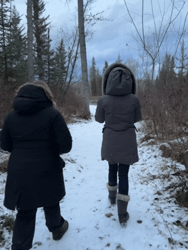 two women are walking down a snowy path with trees in the background