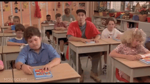 a group of children are sitting at their desks in a classroom with a man in a red shirt standing behind them .