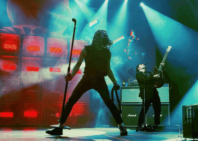 a man singing into a microphone while a man plays a guitar in front of a wall of marshall amps