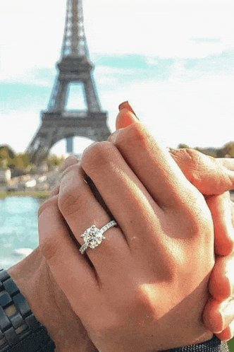 a man and woman are holding hands in front of the eiffel tower . the woman is wearing an engagement ring .