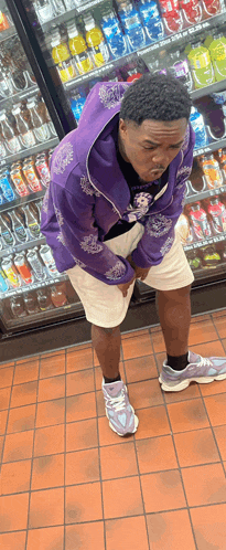 a man in a purple jacket and white shorts is standing in front of a vending machine filled with sodas .