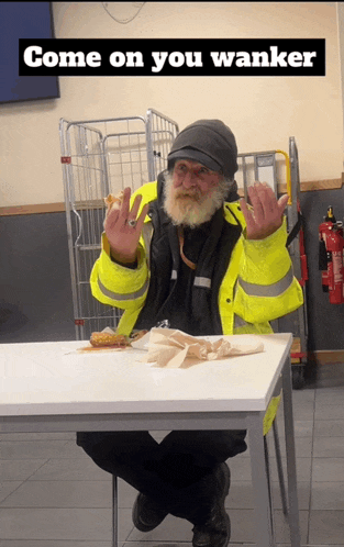 a man with a beard is sitting at a table with a sandwich and giving the middle finger