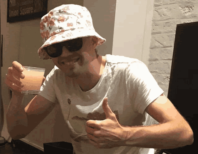 a man wearing a bucket hat and sunglasses holds a drink in front of a sign that says union square