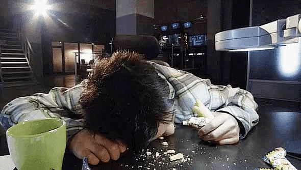 a man is laying on a desk with his head on the table and a green cup in front of him