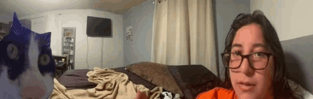 a woman wearing glasses is sitting on a bed with a cat in the background .