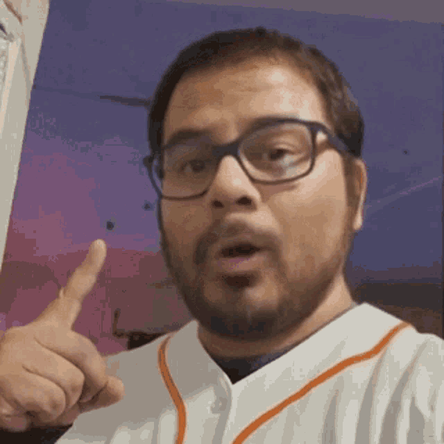 a man with glasses and a beard is wearing a baseball jersey and pointing at the camera .